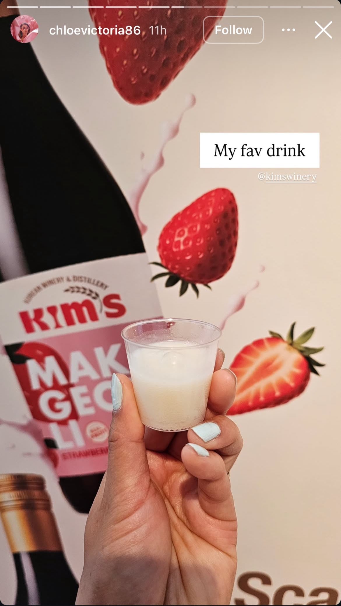 Hand holding a small cup of Kim's Makgeolli against a background of Kim's Strawberry Makgeolli with strawberries floating around.