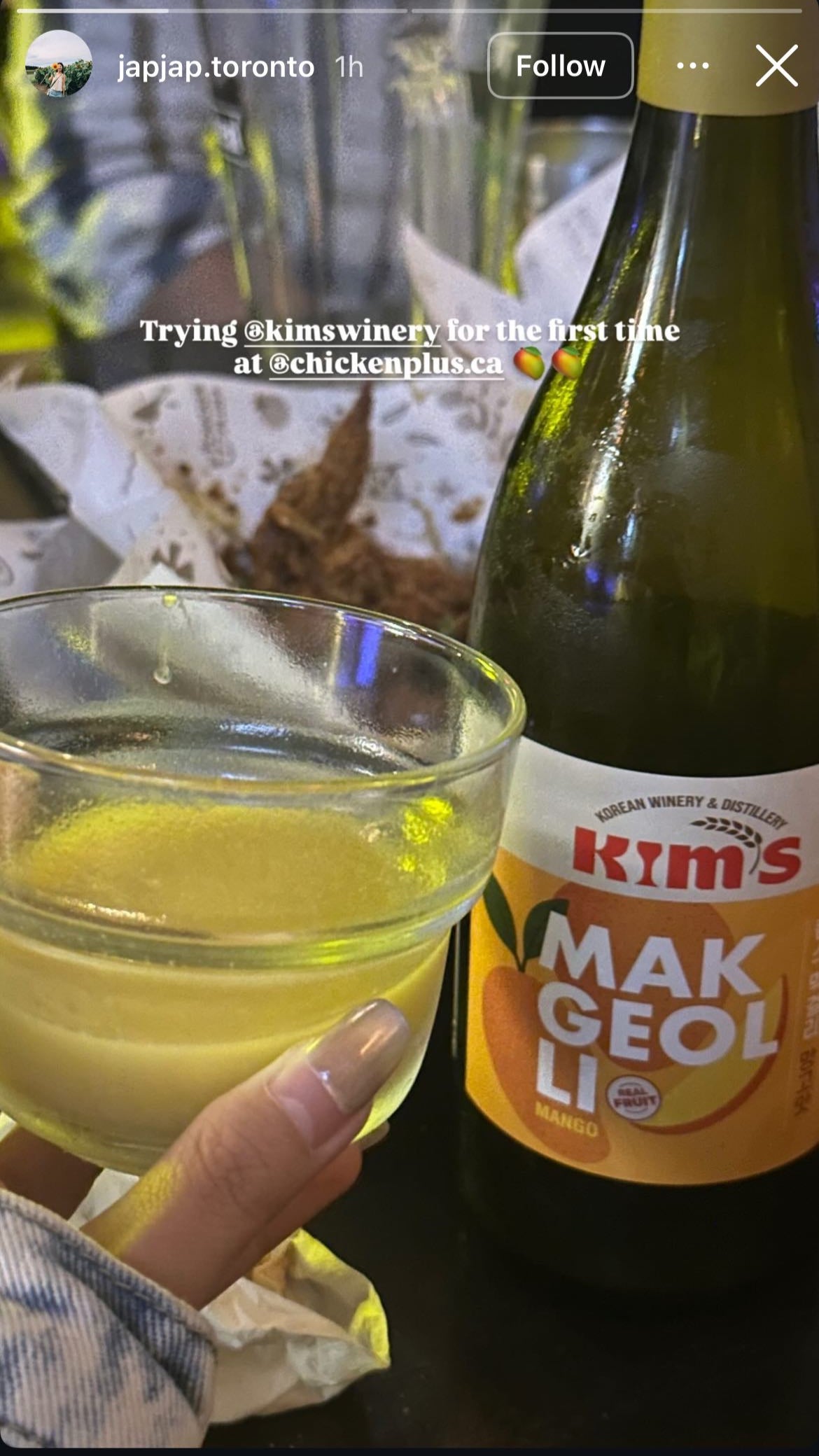 Bottle of Kim's Makgeolli Mango with a glass of yellow mango makgeolli with a background of korean fried chicken on a table.