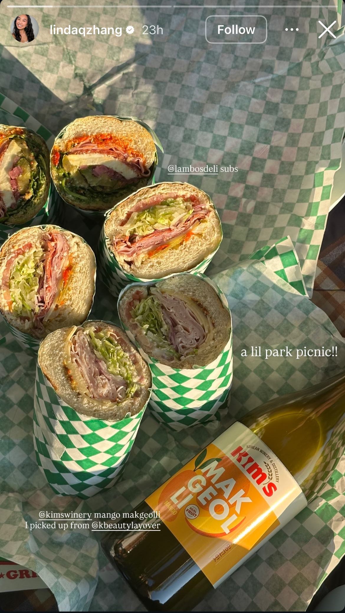 Six deli sub sandwiches with a bottle of Kim's Makgeolli Mango laying on a picnic blanket.