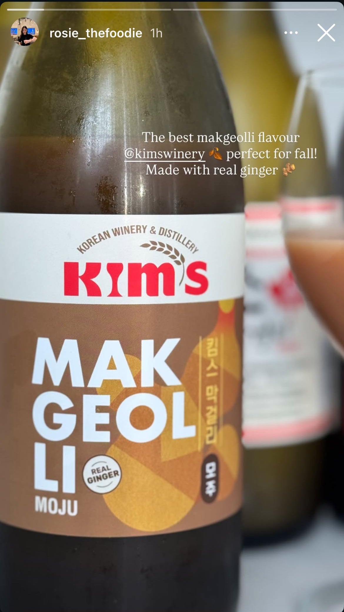 A bottle of Kim's Makgeolli Moju with a wine glass of ginger cinnamon coloured liquid against a blurred background.