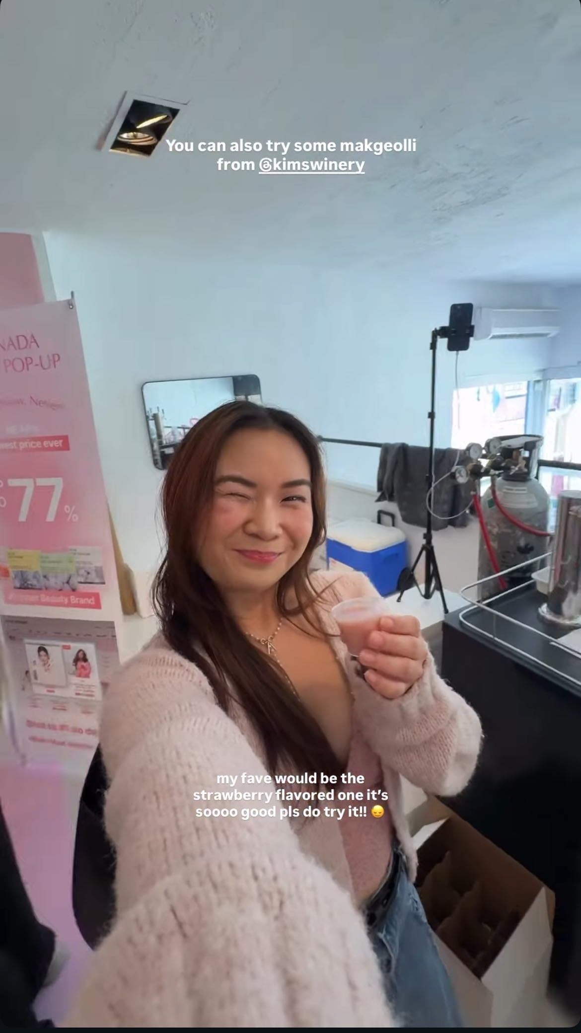 Woman in a pink sweater standing in a store sipping on Kim's Strawberry Makgeolli with a caption recommending the strawberry flavored one is soooo good.